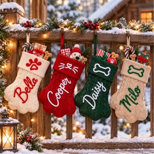 May include: Four paw-shaped Christmas stockings in cream, red, and dark green knit fabric, each featuring a personalized name embroidered in script, a cuff with a paw print or bone motif, and filled with small gifts, candy canes, and plush toys.