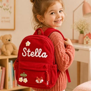 Puede incluir: Mochila de pana roja con el nombre "Stella" en escritura blanca. La mochila tiene un bolsillo frontal con un aplique de zorro y flores. Una mochila roja peque&ntilde;a para ni&ntilde;os.