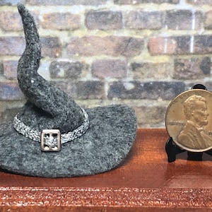 May include: A miniature gray witch hat with a silver band and buckle, next to a 1956 penny displayed on a small black stand. The hat is made of felt and has a curved, pointed top. The penny is bronze-colored.