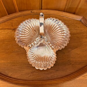 May include: A silver-toned metal serving dish featuring three distinct shell-shaped compartments arranged in a clover-like pattern. The dish includes a central vertical handle that rises from the middle, connecting the three scalloped sections.