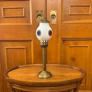 Peut inclure: Une lampe ancienne en verre blanc avec des détails en laiton, ornée d'ornements rouges et bleus. La lampe est posée sur une table d'appoint ovale en bois avec des pieds décoratifs, devant une porte en bois.
