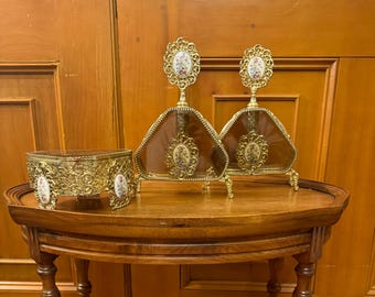 May include: Two gold-toned perfume bottles with floral designs on the stoppers and glass bodies. The bottles are on a wooden table with a gold-toned decorative stand.