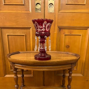 May include: A ruby red glass vase with etched floral designs and hanging crystal accents sits on a small, oval wooden side table. The table has a lower shelf and ornate, turned legs, showcasing a vintage aesthetic.