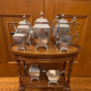 May include: A silver tea set comprising a teapot, creamer, sugar bowl, and two serving cups. The set is displayed on a wooden table with a handwritten note on the bottom shelf.