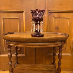 May include: A deep red crystal vase with hanging crystal droplets is displayed on a wooden side table. The vase features an etched pattern and a decorative edge. The oval table has two tiers and turned legs, exuding a vintage charm, with a classic design.