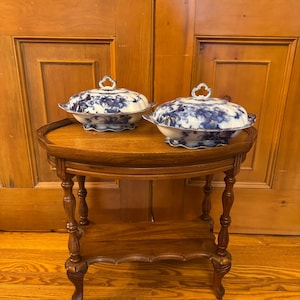 Peut inclure: Deux plats de service en porcelaine bleu et blanc avec couvercles posés sur une petite table d'appoint en bois antique. Les plats présentent un motif floral et des poignées ornées. La table a un plateau rond et des pieds sculptés, et est en bois foncé.