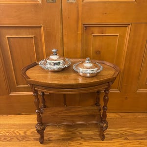 May include: A small, antique wooden side table with two silver serving dishes. The table has an oval top and a lower shelf, supported by ornate, turned legs. The silver dishes have decorative rims and lids, suggesting a formal dining setting.