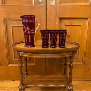 Peut inclure: Une carafe en verre rouge profond avec une anse transparente et quatre verres assortis. Chaque pièce présente des motifs gravés blancs représentant des oiseaux et des fleurs. L'ensemble est disposé sur une table en bois avec une étagère.
