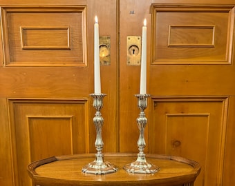 May include: Two silver candle holders with white candles on a wooden table. The candle holders have a decorative design and are in a traditional style.