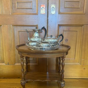 May include: A silver tea set, including a teapot, sugar bowl, and creamer, is presented on a decorative silver tray. The set is displayed on a small, round wooden side table with ornate legs. The background features a pair of wooden doors.