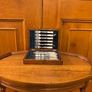 May include: A wooden box containing a set of silverware, including forks and knives, presented on a blue velvet lining. The box is placed on a round wooden table, with a wooden door in the background.