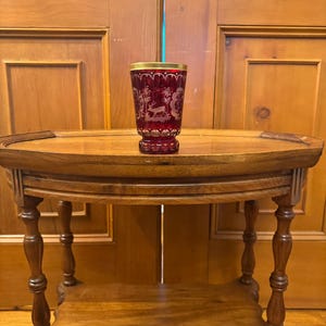 May include: A ruby red glass with a gold rim and etched deer design sits on a wooden table with a lower shelf.