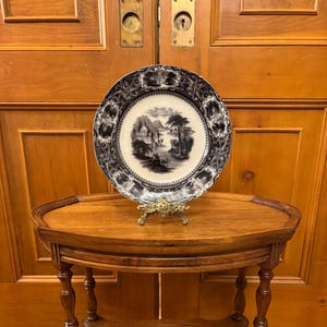 May include: A black and white transferware plate featuring a scenic landscape design. The plate is displayed on a wooden stand.