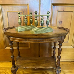 May include: Two antique brass fireplace andirons with a decorative design on a wooden table with a shelf. The table has turned legs and a scalloped edge.