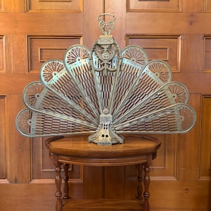 May include: An antique brass fireplace screen in a fan shape, with intricate cut-out designs. The screen is displayed on a small, round wooden table with turned legs, against a wooden door background.