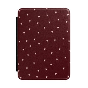 Puede incluir: Una funda rectangular para lector electrónico con una superficie texturizada de color burdeos oscuro, cubierta con un patrón repetitivo de pequeños corazones de color rosa claro. La funda tiene esquinas redondeadas y un lomo negro.