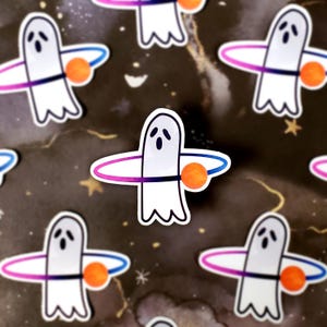 May include: A set of eight white ghost stickers with a pink and blue ring around them and an orange circle inside the ring. The stickers are on a dark brown background with gold and white speckles.