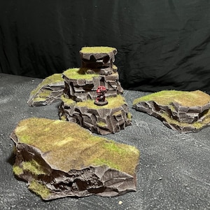 May include: Four miniature rocky terrain pieces for tabletop gaming, painted in brown and green with a small red figure on the tallest piece.
