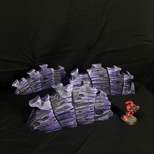 May include: Three purple and grey 3D printed rock formations, resembling a jagged cliff face, with a small red plastic miniature figure standing in front of them.