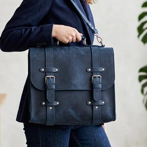 May include: A dark blue leather satchel bag featuring two vertical straps with silver-toned metal buckles, a top handle, and a detachable shoulder strap. The bag has a rectangular flap closure and a smooth, matte finish on the exterior surface.