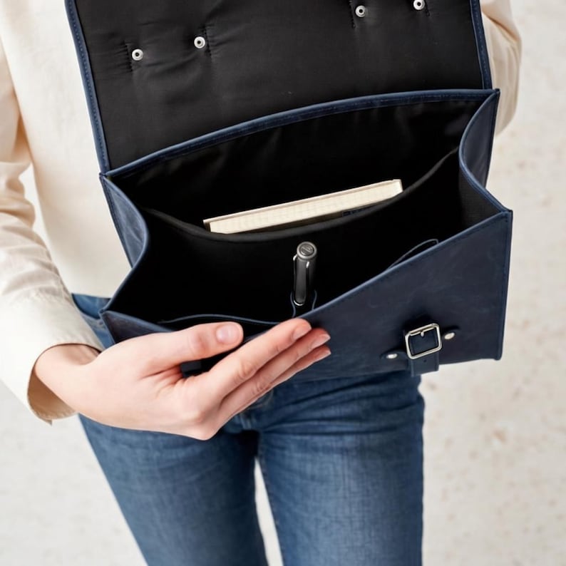 May include: A dark blue satchel with a flap closure, silver-toned metal buckle, and black interior lining. The open bag contains a rectangular notebook and a silver pen held in a dedicated loop, showcasing a structured design for daily organization.
