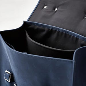 May include: A deep blue leather backpack with a flap top, featuring a silver metal buckle closure on the front. The interior is lined with black fabric and includes a central divider and an open side pocket, with two silver snaps on the flap.