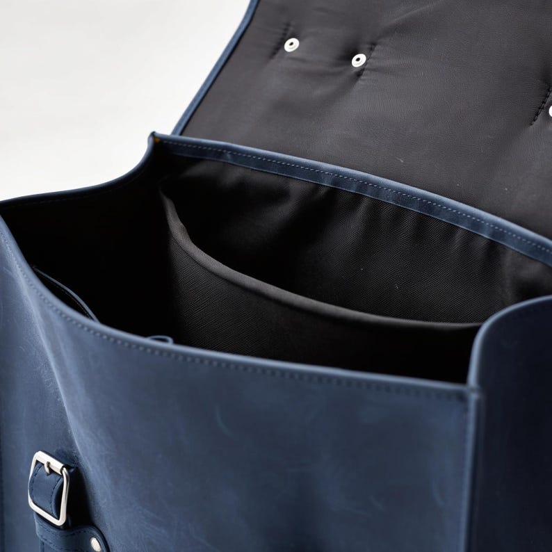 May include: A deep blue leather backpack with a flap top, featuring a silver metal buckle closure on the front. The interior is lined with black fabric and includes a central divider and an open side pocket, with two silver snaps on the flap.