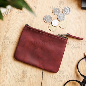 Pu&ograve; includere: Un portamonete in pelle bordeaux con cerniera nera e una piccola linguetta. Diverse monete sono sparse vicino al portamonete su una superficie di legno. Il portamonete &egrave; rettangolare e sembra vuoto. Il marchio "MOSPAN Shop" &egrave; stampato.