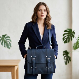 May include: A dark blue leather satchel with a top handle and an adjustable shoulder strap. The bag features a front flap secured by two vertical straps with silver-toned metal buckles and rivets, providing a structured and functional design.