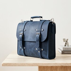 May include: A dark blue satchel with a matte finish, featuring two vertical straps with silver-toned metal buckles, a top handle, and side attachment points for a shoulder strap. The bag has a structured rectangular shape with visible stitching.