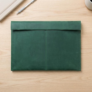 Può includere: Un porta documenti in pelle verde scuro con chiusura a patta. La custodia rettangolare ha una cucitura verticale cucita ed è su una superficie di legno chiaro. Una penna e un taccuino sono visibili sullo sfondo.