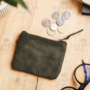 Pu&ograve; includere: Un portamonete in pelle verde scuro con chiusura a cerniera e una piccola linguetta verde. Diverse monete sono sparse intorno al portamonete su una superficie di legno. Il portamonete &egrave; rettangolare.