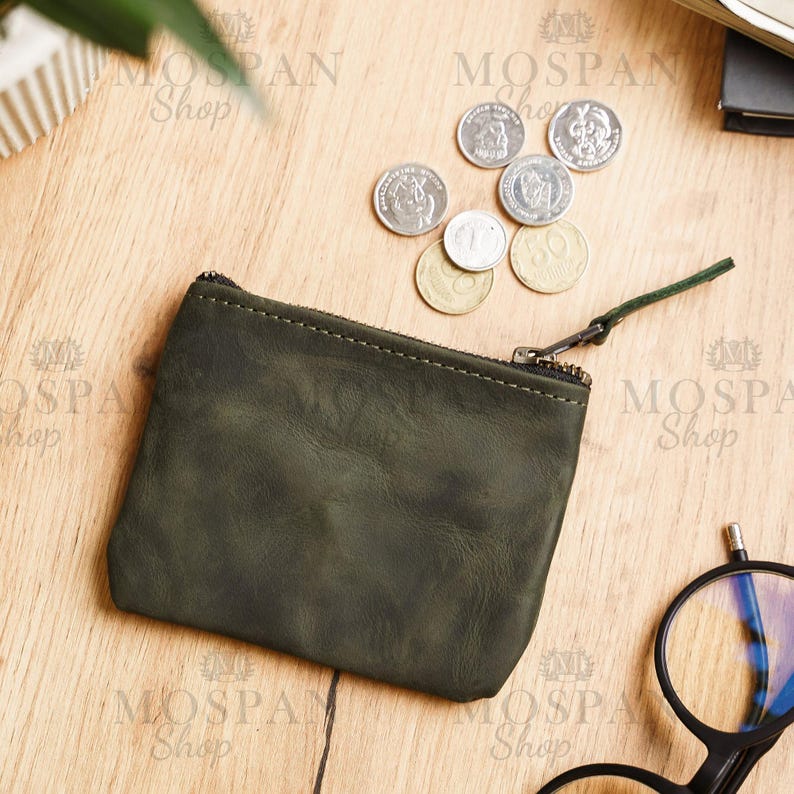Pu&ograve; includere: Un portamonete in pelle verde scuro con chiusura a cerniera e una piccola linguetta verde. Diverse monete sono sparse intorno al portamonete su una superficie di legno. Il portamonete &egrave; rettangolare.