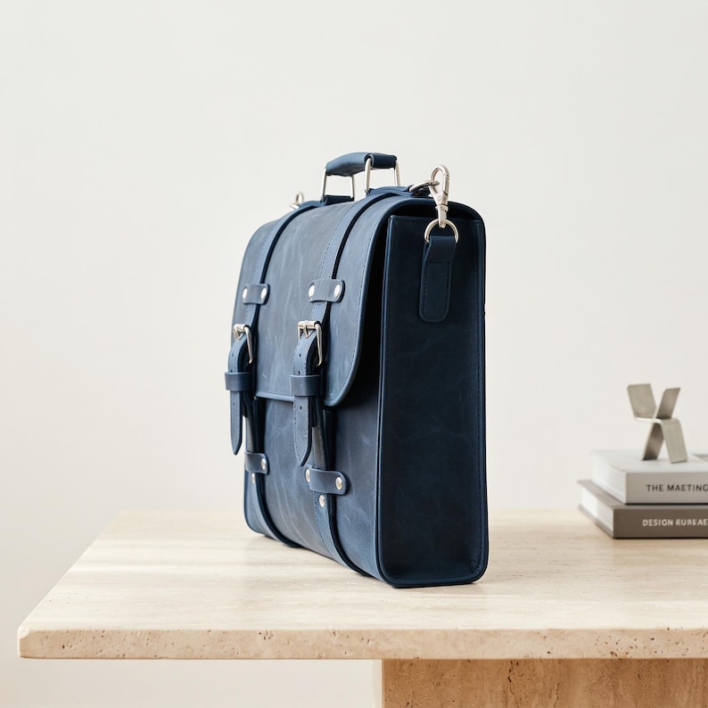 May include: A deep blue leather satchel featuring a top handle, a fold-over flap with two vertical straps, and silver-toned metal buckles. The bag includes a detachable shoulder strap with a silver swivel clasp attached to the side panel.
