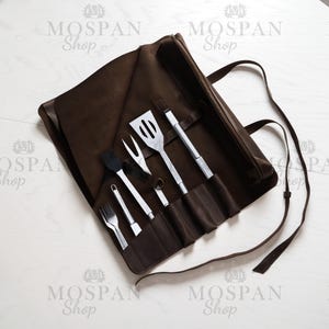 May include: A brown leather roll-up case with a strap, containing a set of six grilling tools: a spatula, tongs, a fork, a basting brush, a small knife, and a small fork. The case is labeled "MOSPAN Shop".