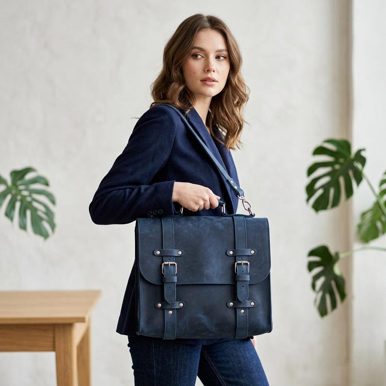 May include: A dark blue leather satchel bag featuring two vertical straps with silver-toned metal buckles, a top handle, and a detachable shoulder strap. The bag has a rectangular flap closure and a smooth, matte finish on the exterior surface.