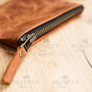 Pu&ograve; includere: Primo piano di un portafoglio in pelle marrone con cerniera in ottone. La cerniera &egrave; parzialmente aperta, rivelando un interno nero. Il portafoglio ha una piccola linguetta in pelle con un rivetto metallico. Sfondo in legno chiaro.