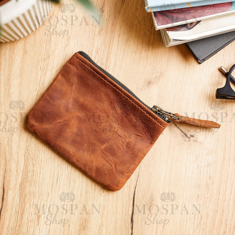 Pu&ograve; includere: Un portamonete in pelle marrone con chiusura a cerniera e tirante in pelle. La borsa &egrave; rettangolare e ha una superficie strutturata. Lo sfondo &egrave; una superficie di legno con altri oggetti.