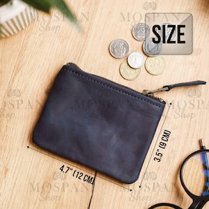 Pu&ograve; includere: Un portamonete in pelle blu navy con chiusura a zip. Il portamonete &egrave; rettangolare, misura 12 cm di larghezza e 9 cm di altezza. Diverse monete sono sparse intorno al portamonete su una superficie di legno. La parola "SIZE" &egrave; in una casella bianca.
