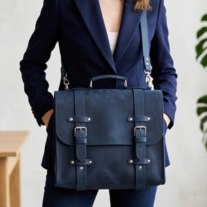 May include: A dark blue leather satchel featuring a top handle, a detachable shoulder strap, and a front flap secured by two vertical straps with metal buckles and silver-toned rivets. The bag has a matte finish and a structured rectangular shape.