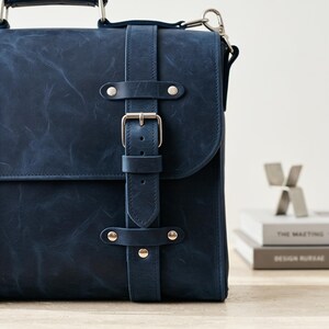 May include: A dark blue leather satchel featuring a vertical strap with a silver-toned metal buckle and rivets. The bag has a top handle and a side attachment ring for a shoulder strap, showcasing a smooth, matte finish with subtle natural texture.