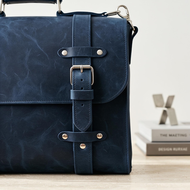 May include: A dark blue leather satchel featuring a vertical strap with a silver-toned metal buckle and rivets. The bag has a top handle and a side attachment ring for a shoulder strap, showcasing a smooth, matte finish with subtle natural texture.