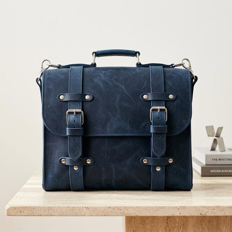 May include: A dark navy blue leather satchel featuring a top handle, a fold-over flap, and two vertical straps secured with silver-toned metal buckles and rivets. The bag has a distressed, matte finish and includes side rings for an optional shoulder strap.