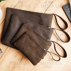 Könnte beinhalten: Vier braune Leder-Reißverschlusstaschen in verschiedenen Größen mit Lederriemen. Die Taschen sind pyramidenförmig auf einer Holzoberfläche angeordnet. Die Taschen sind alle aus dem gleichen braunen Leder gefertigt und haben einen Used-Look.