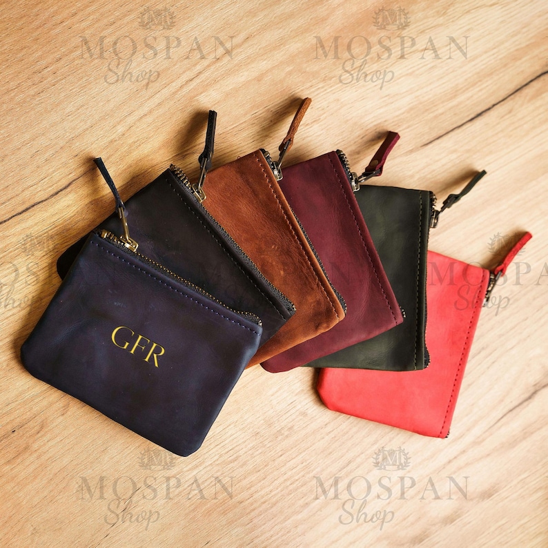 Pu&ograve; includere: Una collezione di portamonete in pelle in vari colori: blu navy con iniziali dorate, marrone, bordeaux, verde scuro e rosso. Ogni portamonete ha una chiusura a zip e un piccolo cinturino da polso. Disposti su una superficie di legno.
