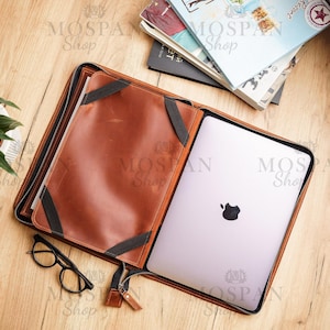 Puede incluir: Una funda para portátil de cuero marrón con cierre de cremallera y un portátil negro en su interior. La funda tiene una solapa con cremallera y una correa negra.