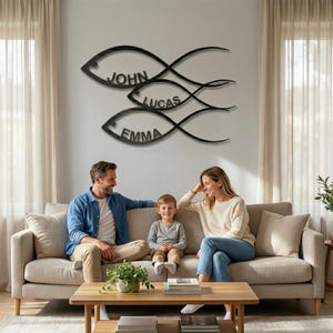 May include: Black metal wall art featuring three fish shapes, each with a name: John, Lucas, and Emma. The artwork is mounted on a light gray wall above a family sitting on a beige sofa. The family is sitting in front of a wooden coffee table.