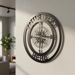 Może przedstawiać: Okrągła, ciemna metalowa dekoracja ścienna z motywem kompasu. Tekst "YOUR TEXT HERE" jest wycięty na zewnętrznej krawędzi. Róża wiatrów jest wyśrodkowana, ze wskaźnikami kierunków. Zamontowana na białej ścianie.