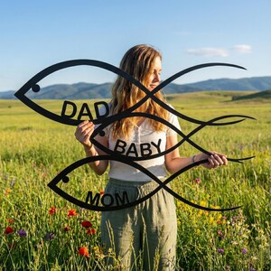 May include: Black metal fish family wall art with the words "DAD", "BABY", and "MOM". The artwork is held in front of a person in a field of wildflowers and tall grass.