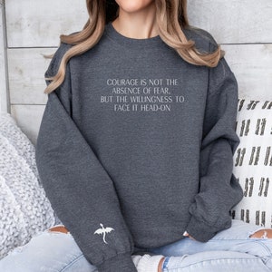 May include: A dark gray sweatshirt with a white quote that reads "Courage is not the absence of fear, but the willingness to face it head-on". A white dragon is embroidered on the left sleeve.